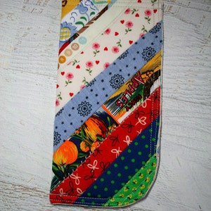 May include: A colorful patchwork fabric eyeglass case with a diagonal pattern. The case is made of various fabrics with different patterns, including floral, polka dots, and stripes. The case is sewn together with a white thread.