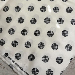 May include: A white fabric featuring a repeating pattern of black circular motifs with a grid-like texture. The selvage edge displays the printed text: andover created by Alison Glass for Andover.