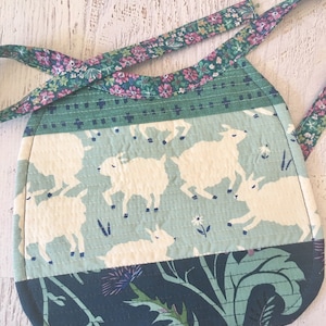 May include: A green, blue, and white baby bib with a floral print on the straps and a pattern of white goats on a light blue background. The bib has a dark blue bottom with a green and white floral print.