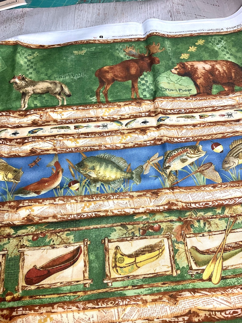 May include: A green fabric with a woodland theme. The fabric features a moose, a wolf, a bear, and a variety of fish. The fabric also includes the text "Iron Wall" and "Brown Bear".