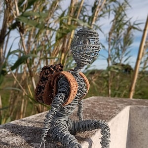 Pode incluir: Escultura de arame de uma pessoa sentada com uma mochila. A figura é feita de arame cinzento, com uma cabeça esférica e um corpo formado por arame torcido. A mochila é de cortiça. A escultura está ao ar livre, com plantas verdes altas ao fundo.