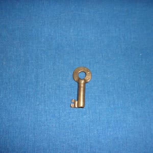 May include: A small, antique-style brass key with a circular head and a rectangular bit. The key is set against a blue background, highlighting its aged appearance and intricate design. The key's details suggest it may be a vintage or decorative item.