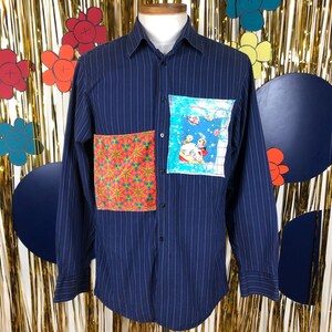 Peut inclure: Une chemise à manches longues boutonnée bleu marine à rayures blanches. La chemise a deux patchs en tissu cousus sur le devant. Un patch est un motif géométrique en rouge, orange et bleu. L'autre patch est une illustration colorée d'une personne dans un vaisseau spatial.