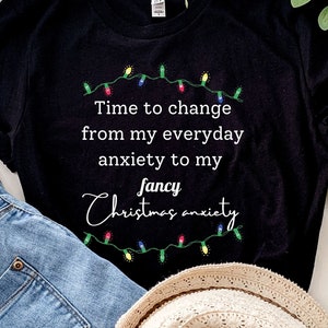 Può includere: Maglietta nera con testo bianco che recita "Time to change from my everyday anxiety to my fancy Christmas anxiety". Il testo è circondato da una stringa di luci natalizie colorate.