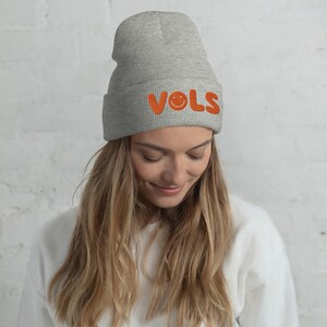 May include: A gray beanie with the word "VOLS" embroidered in orange, along with a smiling face. The beanie is made of a soft, knitted material.
