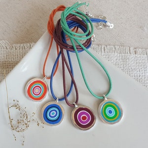 Puede incluir: Cuatro collares con colgantes circulares de colores. Los collares están hechos de cordones de diferentes colores y los colgantes son de plata con círculos concéntricos de rojo, azul, verde y morado.