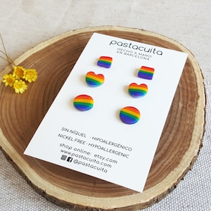 Puede incluir: Una tarjeta blanca muestra seis pares de pendientes de colores del arcoíris en varias formas: cuadrados, corazones y círculos. La tarjeta incluye texto en español e inglés, incluyendo "NICKEL FREE - HYPOALERGÉNICO" y detalles de la tienda. Los pendientes se presentan sobre una rodaja de madera.