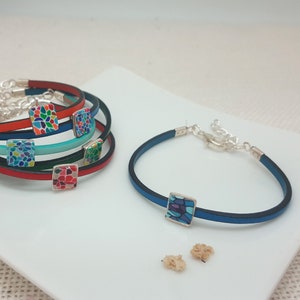 Pode incluir: Um conjunto de cinco pulseiras coloridas com pingentes quadrados de mosaico. As pulseiras são de couro e têm fechos prateados. Os pingentes são de mosaico e apresentam padrões diferentes.