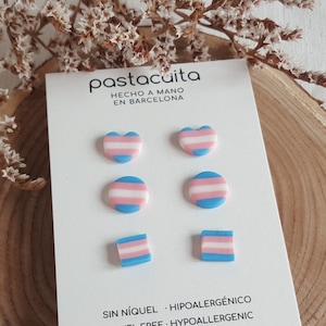 Op de afbeelding: Een set van zes transgender pride-vlag oorbellen in drie verschillende vormen: hart, cirkel en vierkant. De oorbellen zijn gemaakt van polymer clay en zijn nikkelvrij en hypoallergeen. De tekst op de kaart luidt: "pastacuita, Hecho a mano en Barcelona, Sin níquel, Hipoalergénico, Nickel Free, Hypoallergenic, shop online: etsy.com, www.pastacuita.com, @pastacuita".