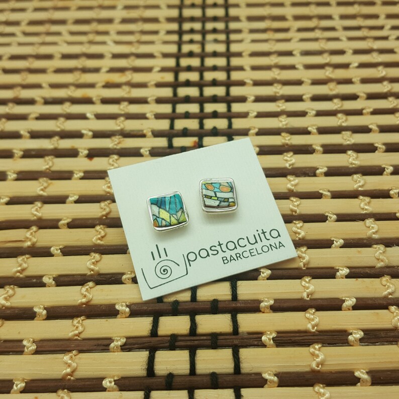 May include: A pair of square-shaped earrings with a colorful mosaic design. The earrings are silver-toned and have a white background with blue, green, and yellow accents. The earrings are displayed on a white card with the text "pastacuita BARCELONA".