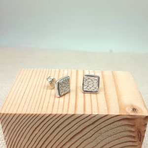 May include: A pair of silver stud earrings with white square-shaped ceramic inlays featuring a geometric pattern.
