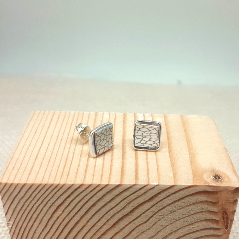 May include: A pair of silver stud earrings with white square-shaped ceramic inlays featuring a geometric pattern.