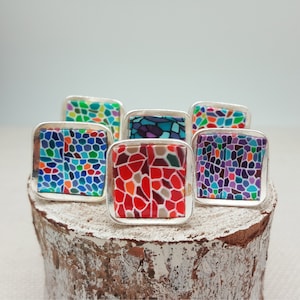 Könnte beinhalten: Sechs silberne Ringe mit bunten Mosaikmustern. Die Ringe zeigen eine Vielzahl von Farben, darunter Rot, Blau, Grün, Lila und Gelb. Die Ringe sind in einer Reihe auf einer Holzoberfläche angeordnet.