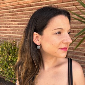 Puede incluir: Una mujer con un top negro de tirantes y pendientes cuadrados plateados con una piedra azul en el centro.