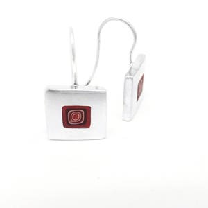 Puede incluir: Un par de pendientes de plata con un dise&ntilde;o cuadrado. Cada pendiente presenta un dise&ntilde;o de c&iacute;rculos conc&eacute;ntricos rojo y blanco dentro del cuadrado.