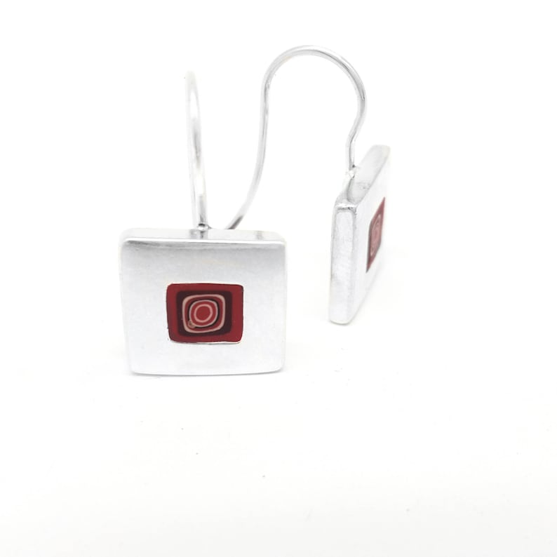 Puede incluir: Un par de pendientes de plata con un dise&ntilde;o cuadrado. Cada pendiente presenta un dise&ntilde;o de c&iacute;rculos conc&eacute;ntricos rojo y blanco dentro del cuadrado.