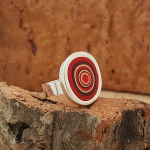 May include: A silver ring with a circular, layered design. The ring's face features concentric circles in shades of red, cream, and brown, set within a silver border. The ring is displayed on a piece of wood, showcasing its design.