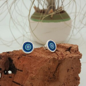Puede incluir: Pendientes de botón plateados con un diseño circular. Los pendientes presentan círculos concéntricos en tonos azules, turquesas y morados, enmarcados por un borde plateado. Los pendientes se presentan sobre un trozo de madera rojizo-marrón.