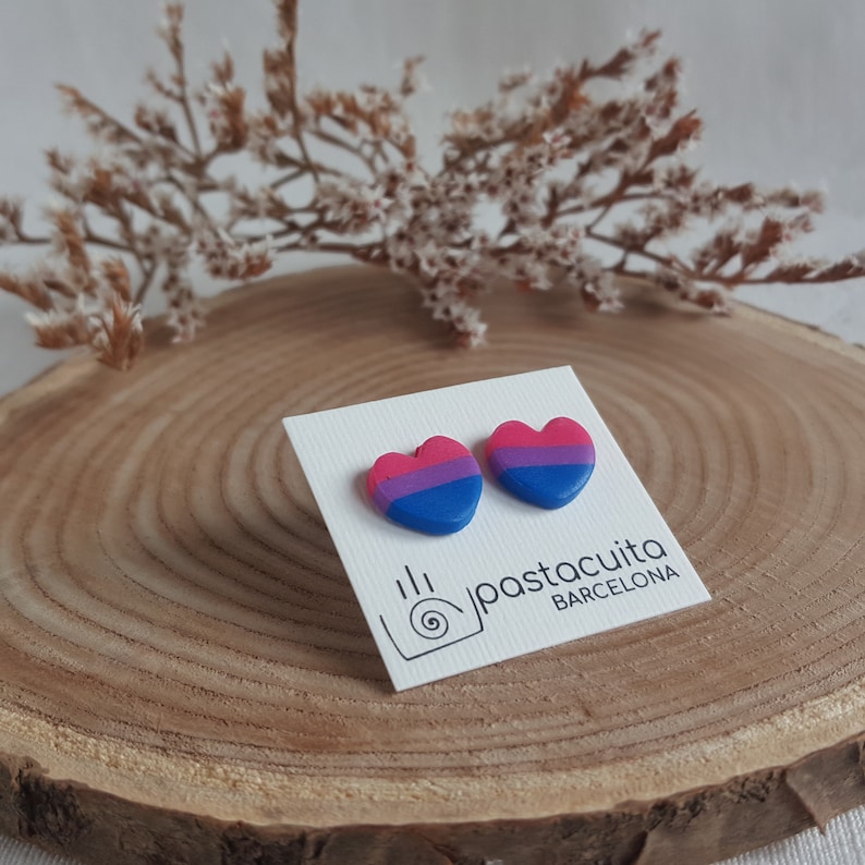 May include: A pair of heart-shaped earrings with the bisexual pride flag colors. The earrings are on a white card with the text "pastacuita BARCELONA".