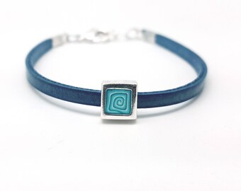 Puede incluir: Un brazalete de cuero azul con un dije cuadrado plateado con un diseño de espiral turquesa.