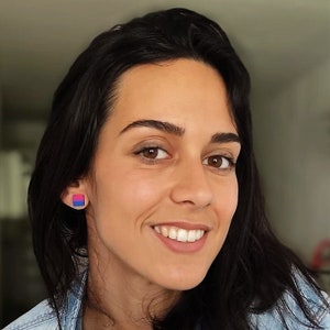 May include: A woman with long dark hair is smiling at the camera. She is wearing a blue denim shirt and has a small square earring in her left ear. The earring is blue, pink, and purple.