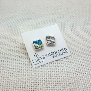 May include: A pair of silver stud earrings with square ceramic tiles featuring colorful geometric patterns. The tiles are blue, green, yellow, orange, and white. The earrings are displayed on a white card with the text "pastacuita BARCELONA".