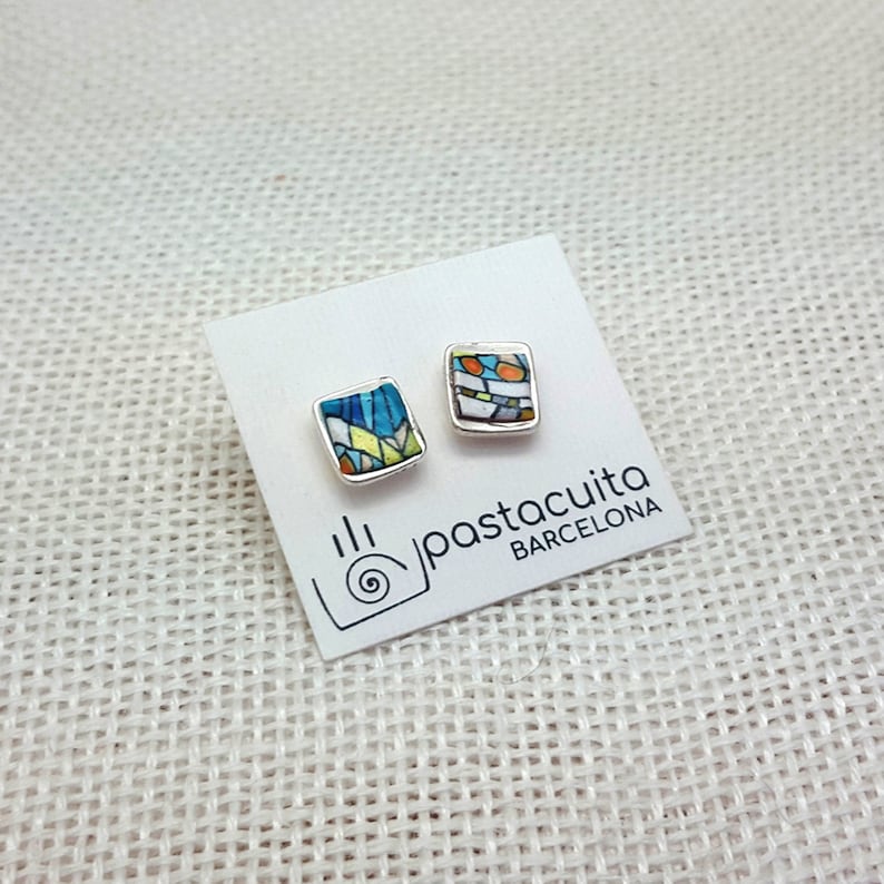 May include: A pair of silver stud earrings with square ceramic tiles featuring colorful geometric patterns. The tiles are blue, green, yellow, orange, and white. The earrings are displayed on a white card with the text "pastacuita BARCELONA".