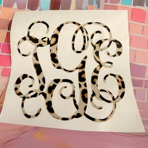 May include: A decorative vinyl decal featuring a three-letter monogram in a cursive script font. The letters are filled with a repeating leopard print pattern consisting of black, brown, and tan spots on a light beige background.