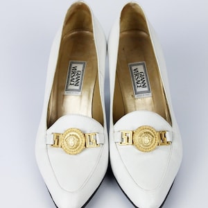 May include: White leather loafers with gold-tone Medusa head detailing. The shoes have a pointed toe and a low heel. The insole features the "Gianni Versace" label. The shoes are a classic style.
