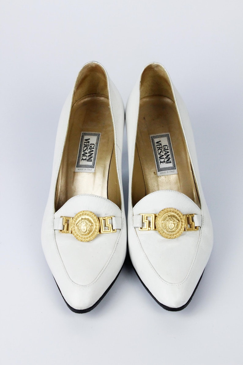 May include: White leather loafers with gold-tone Medusa head detailing. The shoes have a pointed toe and a low heel. The insole features the "Gianni Versace" label. The shoes are a classic style.