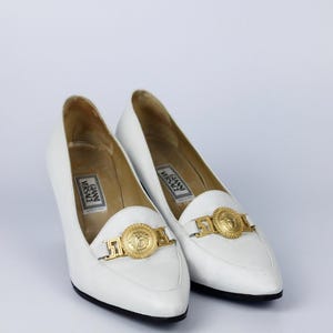 May include: A pair of white leather heels with pointed toes. Each shoe features a gold-tone medallion and buckle detail on the vamp. The heels have a low, stacked heel and the brand name "Gianni Versace" is visible inside.