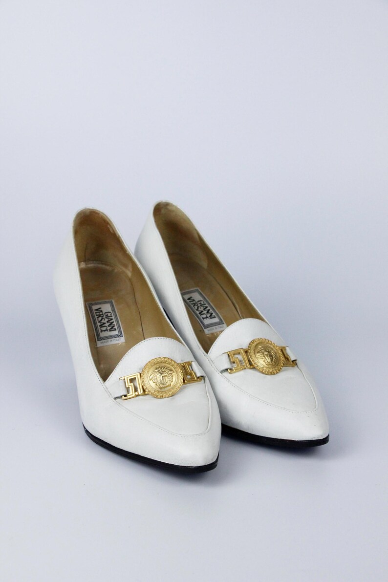 May include: A pair of white leather heels with pointed toes. Each shoe features a gold-tone medallion and buckle detail on the vamp. The heels have a low, stacked heel and the brand name "Gianni Versace" is visible inside.