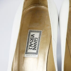 May include: Close-up of white leather high heels. The interior of the shoe is gold-toned, with a label that reads "GIANNI VERSACE MADE IN ITALY". The shoes are set against a white background.