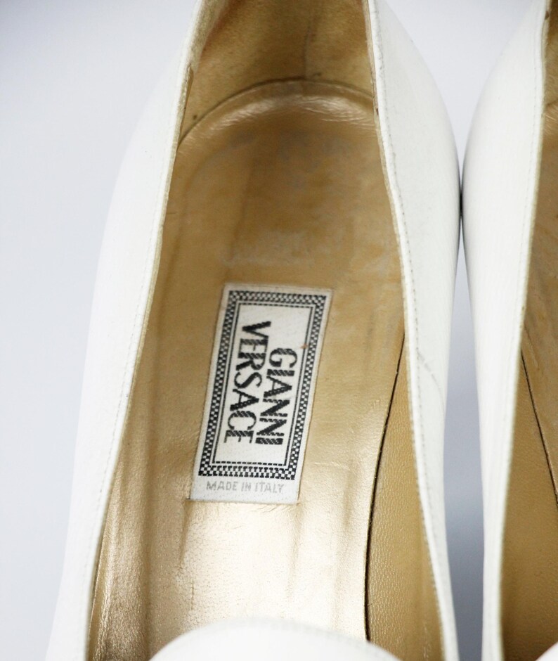 May include: Close-up of white leather high heels. The interior of the shoe is gold-toned, with a label that reads "GIANNI VERSACE MADE IN ITALY". The shoes are set against a white background.