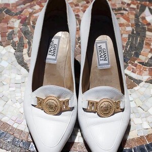 May include: A pair of white leather loafers with a pointed toe and a gold-tone metal emblem. The shoes have a low heel and the label "GIANNI VERSACE" is visible inside.