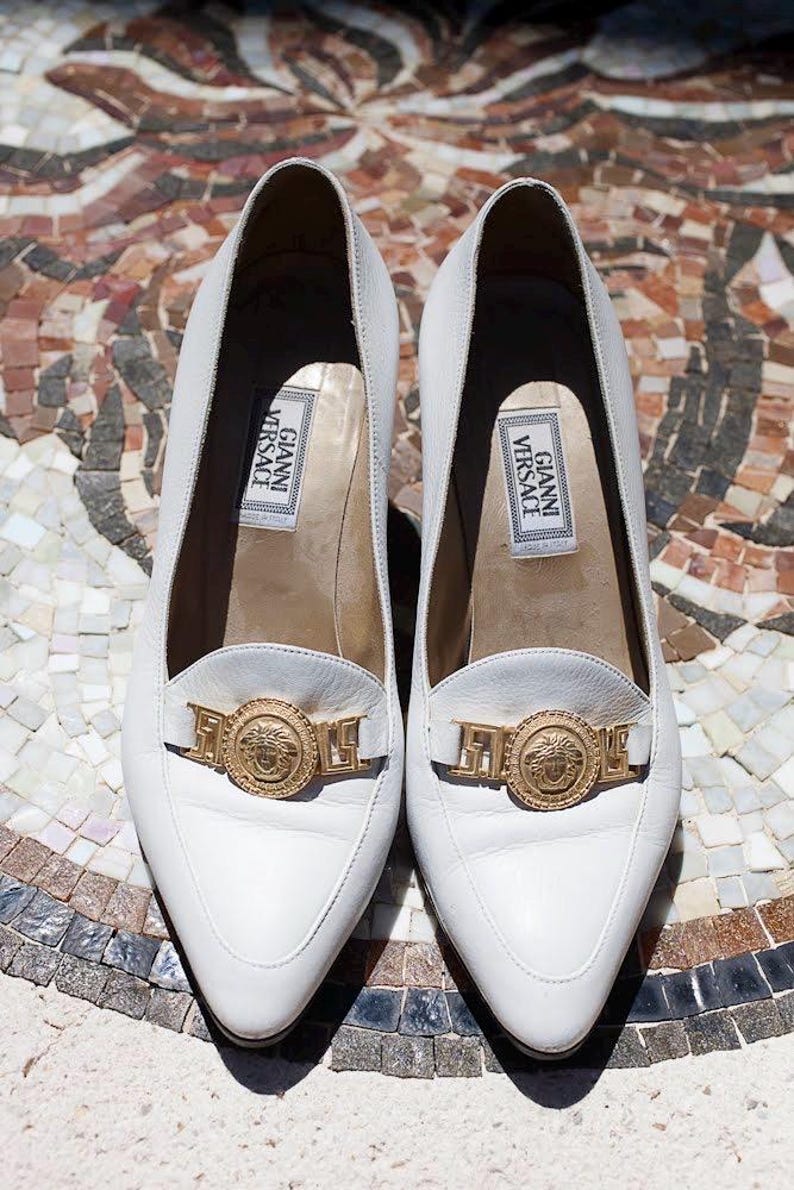 May include: A pair of white leather loafers with a pointed toe and a gold-tone metal emblem. The shoes have a low heel and the label "GIANNI VERSACE" is visible inside.