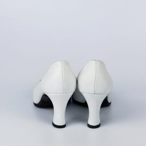 May include: A pair of white leather pumps with a low, curved heel. The shoes have a classic design with a rounded toe and a small heel, approximately 5 cm high. The soles are black.