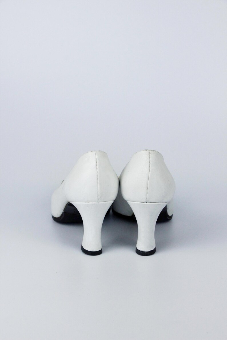 May include: A pair of white leather pumps with a low, curved heel. The shoes have a classic design with a rounded toe and a small heel, approximately 5 cm high. The soles are black.