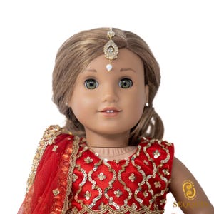 Puede incluir: Una muñeca con un atuendo de estilo indio rojo y dorado con una diadema adornada con joyas. La muñeca tiene el pelo castaño y los ojos azules.