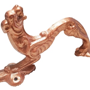 Puede incluir: Un tirador de puerta ornamentado de color cobre con un diseño decorativo y curvo. El tirador presenta una apariencia detallada y esculpida con un patrón de volutas y una cabeza de animal estilizada en la parte superior. El tirador está diseñado para montarse en una puerta.