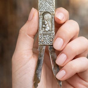 May include: An antique silver key with an intricate design. The key showcases a detailed relief of a squirrel within a decorative frame. The key's blades are extended, revealing their aged metal appearance. The key is held by a person.