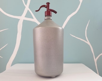 Pode incluir: Uma garrafa de seltzer prateada vintage com uma alavanca e um bico vermelhos. A garrafa tem uma superfície texturizada e uma base arredondada. O fundo é uma parede azul clara com desenhos brancos em forma de galhos.
