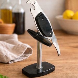 Puede incluir: Un abridor de botellas con forma de pájaro hecho de metal plateado y material negro, montado sobre un poste vertical unido a una base rectangular negra. La figura del pájaro presenta un pico estilizado que funciona como abridor y un mango negro.