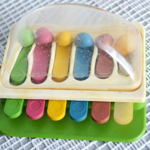 May include: A colorful toy xylophone with a clear plastic cover. The xylophone has a green base and a cream-colored frame. The keys are various colors, including green, pink, yellow, blue, and brown. The mallets are also colorful.