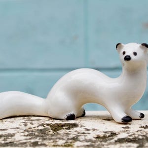 May include: A white ceramic figurine of a weasel or stoat. The figurine is posed in a running position with its tail curled up behind it. The figurine has black eyes and a black nose.