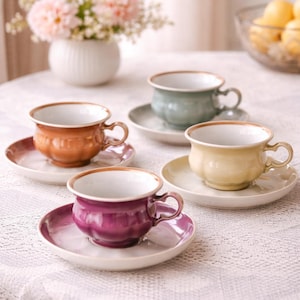 Könnte beinhalten: Vier Keramik-Tassen und Untertassen in Orange-, Grün-, Gelb- und Lilatönen. Jede Tasse hat ein gewelltes Design und einen braunen Rand. Die Untertassen passen zu den Tassenfarben. Im Hintergrund eine Vase mit Blumen und eine Schüssel mit Zitronen.