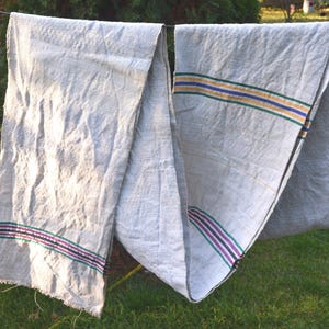 May include: Two light gray linen textiles hanging on a clothesline. Each textile has a striped border in shades of purple, green, and yellow. The textiles are hanging outdoors on a sunny day.