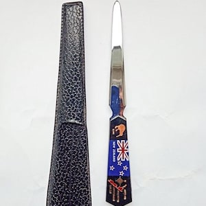 Puede incluir: Un abrecartas con una hoja plateada y un mango con la bandera de Nueva Zelanda, un kiwi y el texto "NEW ZEALAND". Viene con una funda negra texturizada.