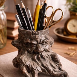 May include: A detailed, bronze-colored desk organizer shaped like a bearded face, holding pencils, pens, and gold scissors. The pencils are black and yellow. The organizer sits on a neutral-colored book.