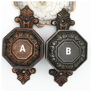 Könnte beinhalten: Zwei antike Schrankgriffe in Bronze- und Silberfarben. Jeder Griff hat eine achteckige Fläche mit strukturiertem Zentrum und aufwendigen floralen Details. Die Griffe sind mit "A" und "B" beschriftet.